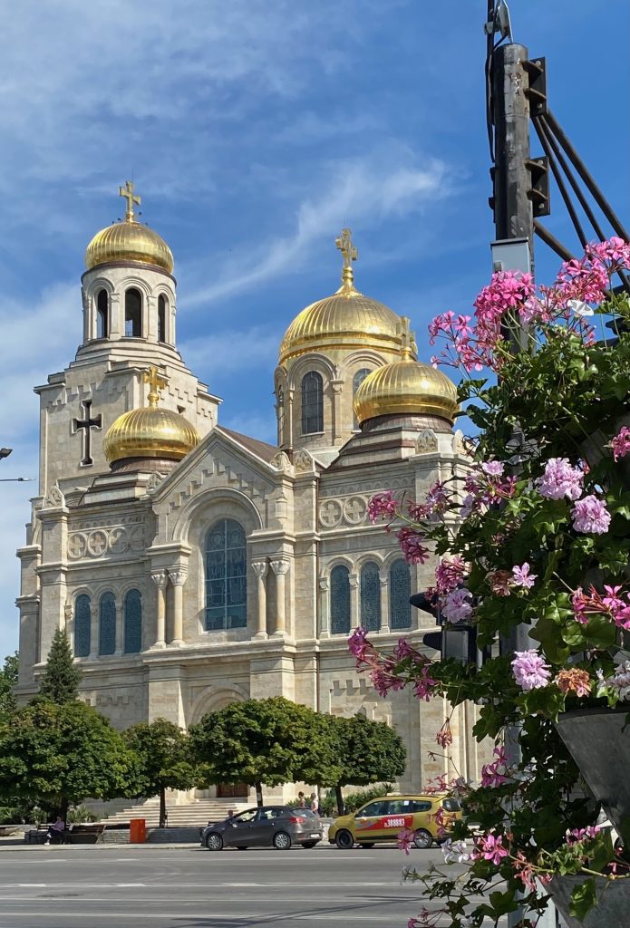 Sandsteinfarbene Kirche mit goldenen Kuppeln in Varna, im Vordergrund rosafarbene Blumen