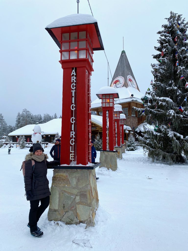 Marianne neben einer Säule mit der Aufschrift "Arctic Circle" am Santa Claus Village bei Rovaniemi in Finnland
