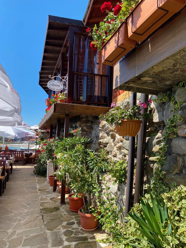 Kleine Gasse in Sosopol in Bulgarien mit Steinmauer, gesäumt von Pflanzkpbeln mit strahlend grünen Blättern und rosafarbenen Blüten, im Hintergrund Tische eines Restaurants