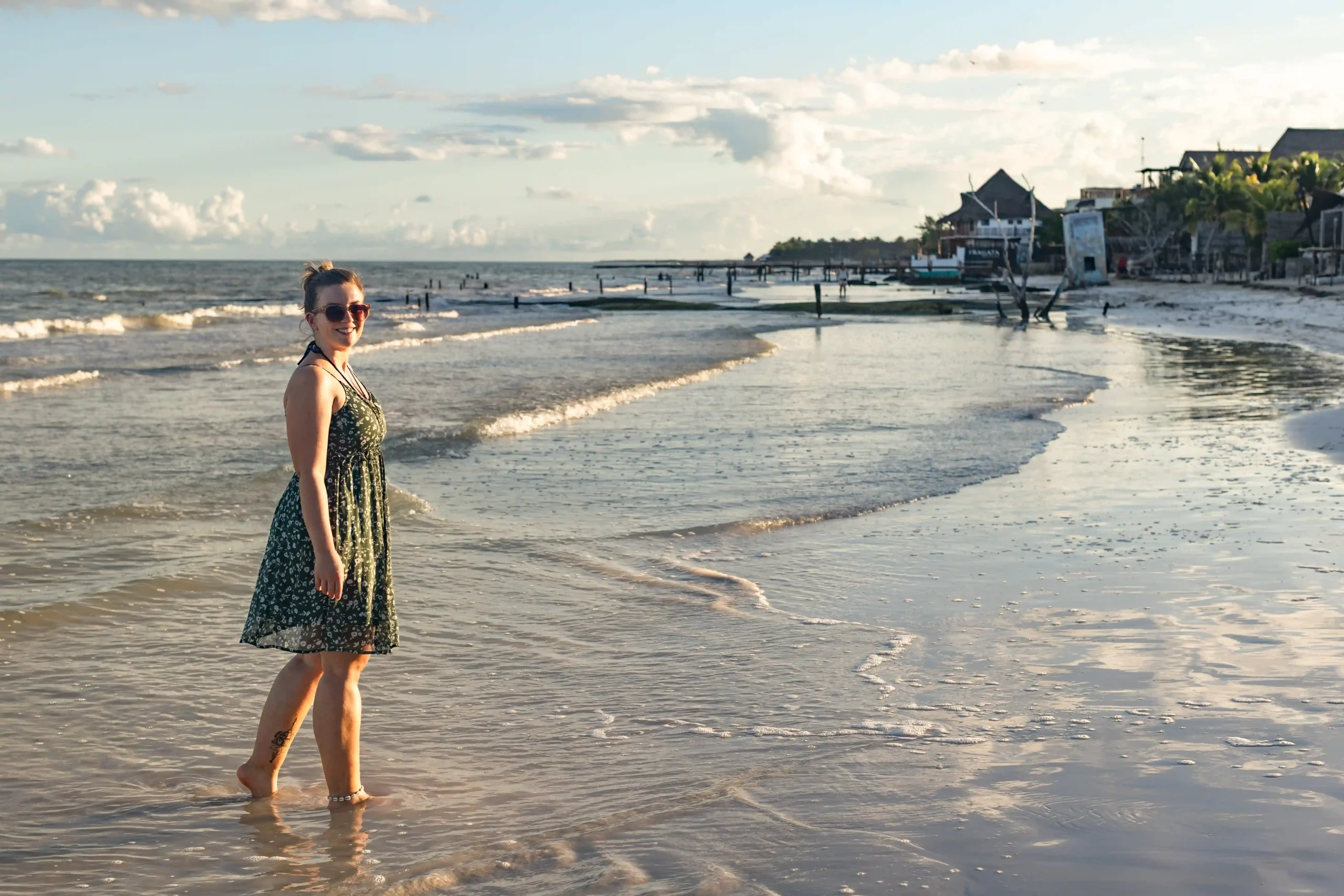 Marianne am Strand von Holbox mit den Füßen im flachen Wasser, im Hintergrund Häuser und Palmen