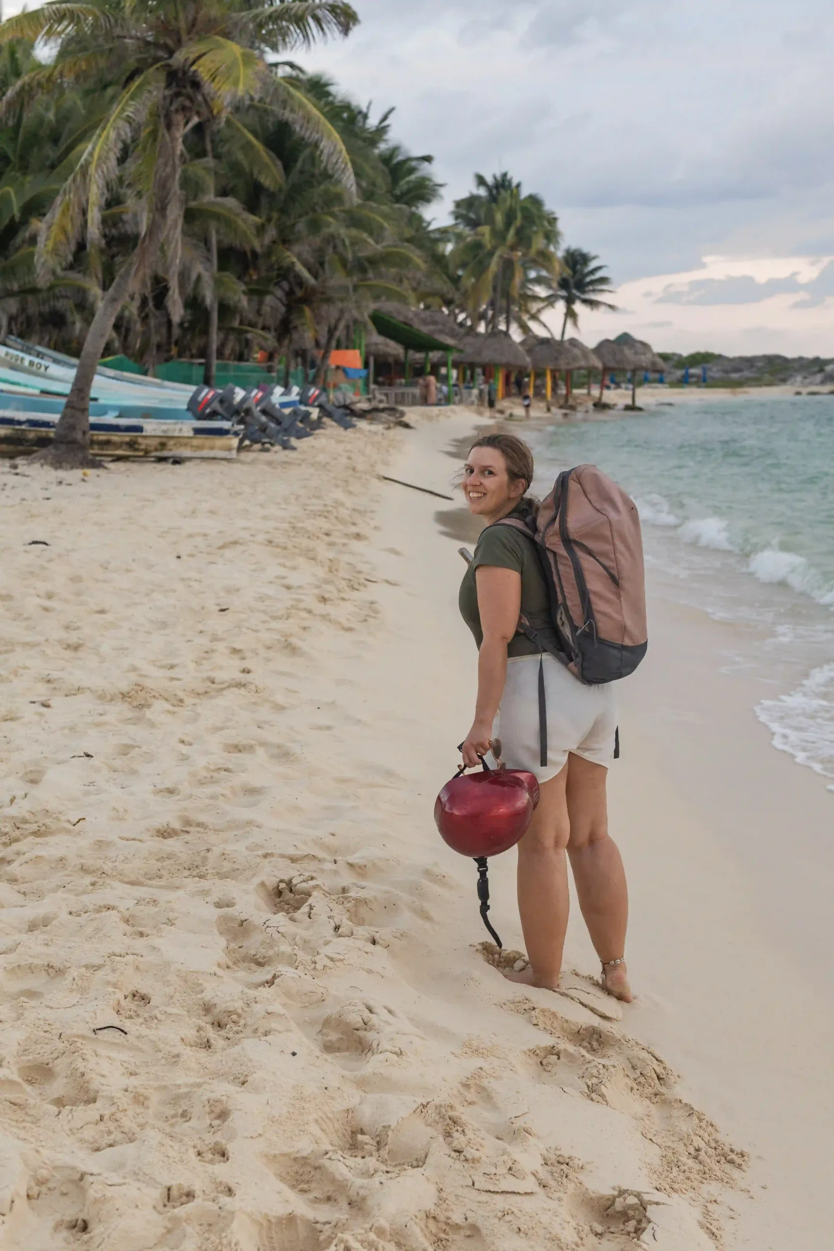 Marianne mit Rucksack und Roller-Helm in der Hand an einem Sandstrand von Cozumel, im Hintergrund Palmen und kleine Boote