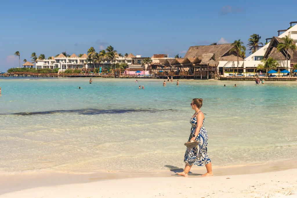 Marianne läuft entlang des Sandstrandes Playa Norte auf Isla Mujeres, dahinter türkisblaues Meer, in dem einige Menschen sind, im Hintergrund Palmen und Häuser von Hotelanlagen